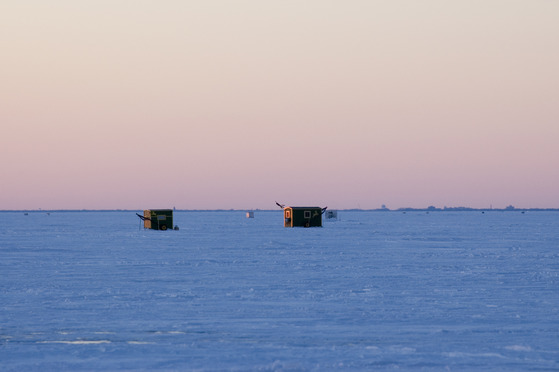Two ice shanties siting on the ice around sunrise or sunset. 