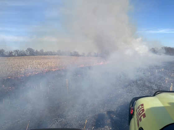 The hood of a fire truck is seen in front of a burning field giving off dark gray smoke. 