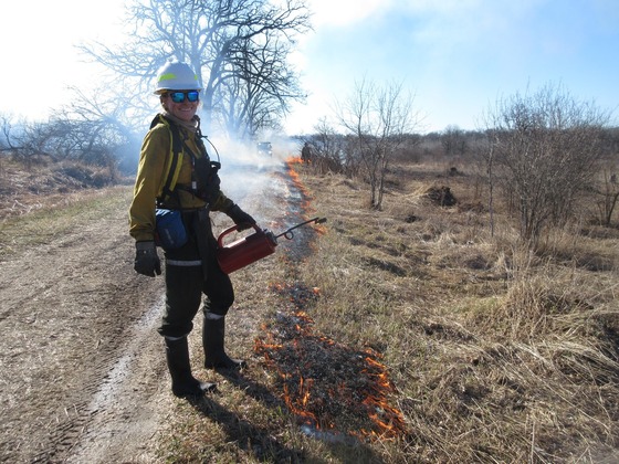 a smiling male with safety gear conducting a prescribed burn in a grassy area