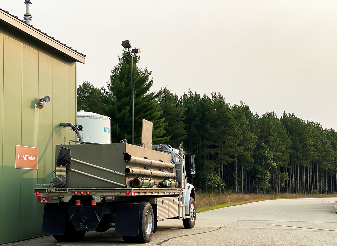 A large beige fish stocking truck fills up with fish from a forest green building with a sign on it reading "Headtank." 