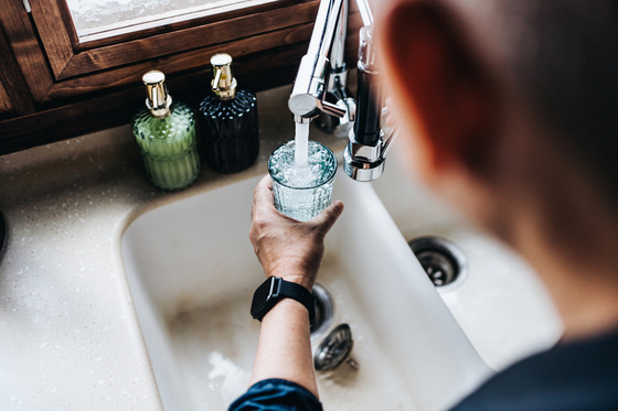 A behind-the-shoulder view of a man pouring himself a glass of water from a kitchen faucet. 