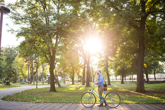 A man in business attire pushes a bike along a cobblestone sidewalk as the sun peeks through green trees in the background. 