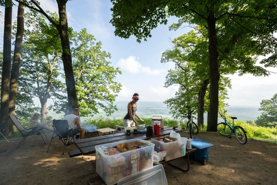 Three people sit around a campfire next to a picnic table while viewing the scenery from an elevated campsite at Wyalusing State Park. 