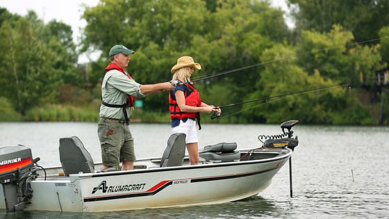 A man and woman wearing life jackets stand in an aluminum boat and fish. 