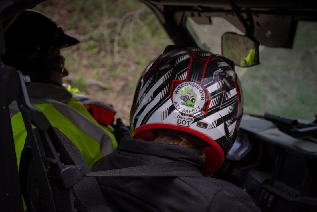 Two people, both wearing helmets, ride in a UTV. The rider on the right has a ATV Safety Graduate sticker displayed on their helmet. 