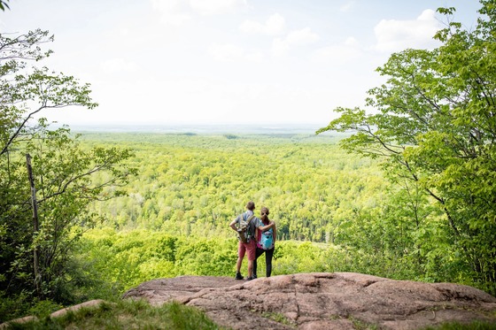 A couple stands on a rocky overlook hundreds of feet above an expansive, green forestscape. 