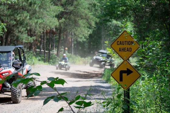 A yellow trail sign reading "Caution Ahead" with a right turn arrow is seen in front of a gravel trail with ATVs riding on it. 