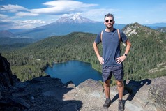 Dan Noreika smiling while standing on a rock overlooking a lake, coniferous forest and Mount Shasta in California.