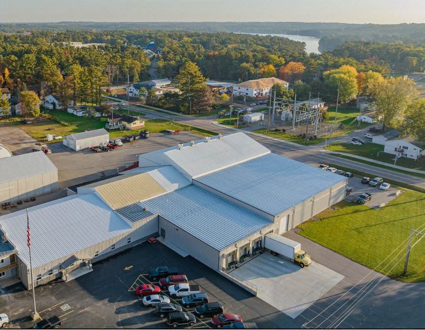 A metal warehouse facility as viewed from a drone. A river lined with tall trees can be seen in the background. 