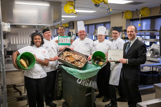 A group of chefs stand together with compost buckets in a commercial kitchen. 