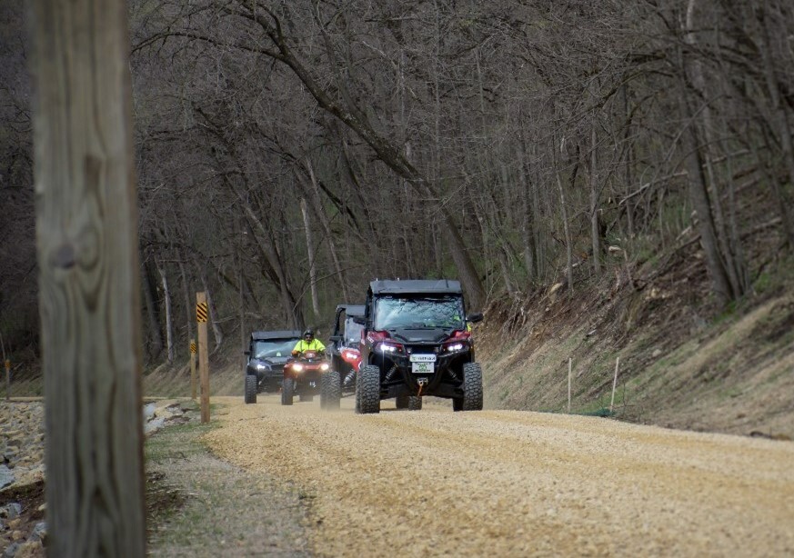 Three UTVs and one ATV ride in a line along a gravel trail. 