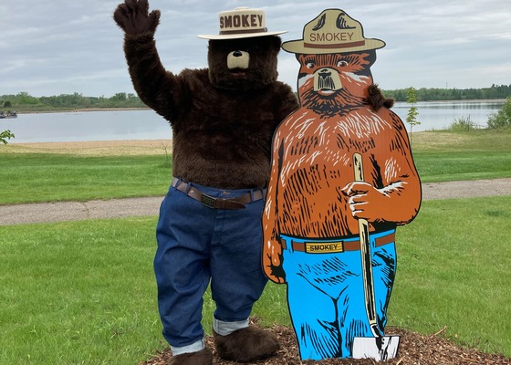 A Smokey Bear mascot waves while posing alongside an animated Smokey Bear cutout with a small lake in the background. 