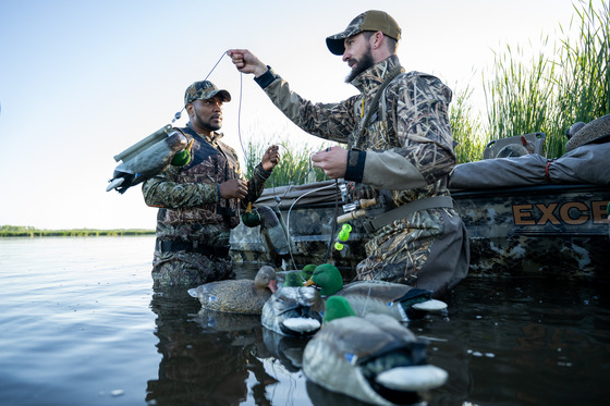 Two individuals in camo wade in the water and set out mallard decoys. 