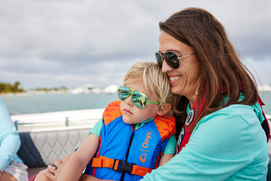 A smiling mother holds a young son on her lap, both wearing life jackets.