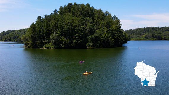 A large, forested island in a blue lake. Five kayakers paddle on the lake. 