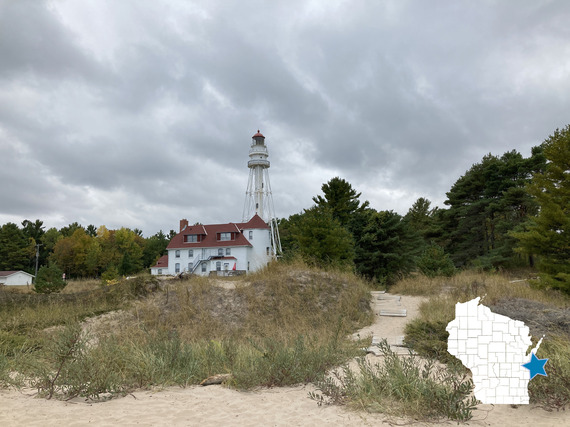 A white lighthouse with a red roof on a sandy beach. Storm clouds are in the sky and a row of trees lines the horizon behind the lighthouse.