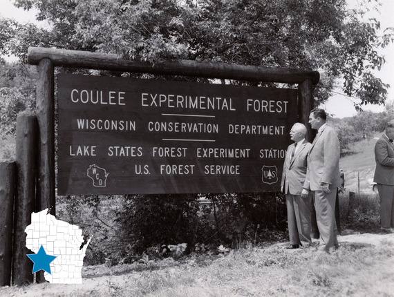 Black and white image of two people stand in front of a large wooden sign that reads "Coulee Experimental State Forest”. 