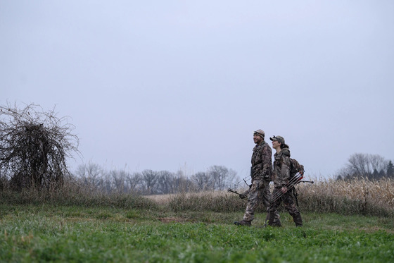 a male and female wearing camouflage walking near grassy area carrrying bows