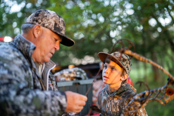 A man in camo holds a bow while a young boy in camo looks on. 