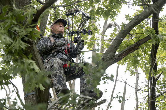 A man wearing all camo sits in a tree stand holding a bow.