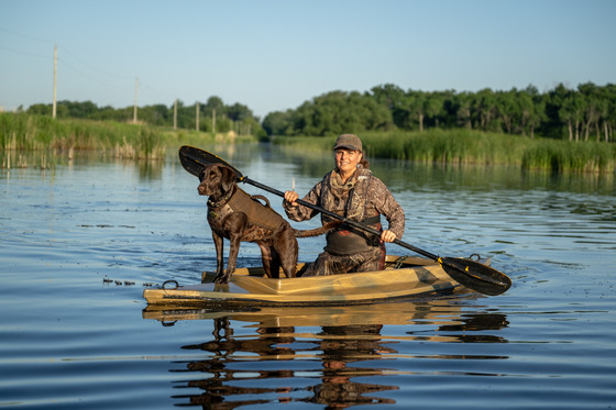 A person in all camo paddles a duck skiff on calm water with a chocolate lab standing attentively on the prow. 