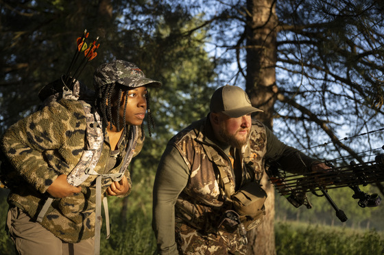 a woman and man wearing camouflage and carrying bows looking at something in the woods