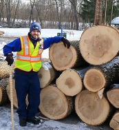 A man in a reflective vest stands next to a large stack of tree logs in a snow-covered yard.