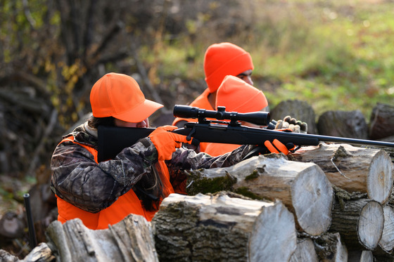 Three people, all wearing blaze orange, kneel behind a large pile of wood while one aims their rifle at a deer out of frame. 