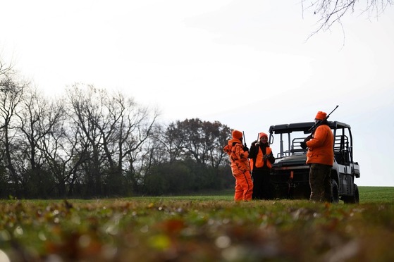 Three individuals in blaze orange clothing hold guns while standing around a UTV in an open field.