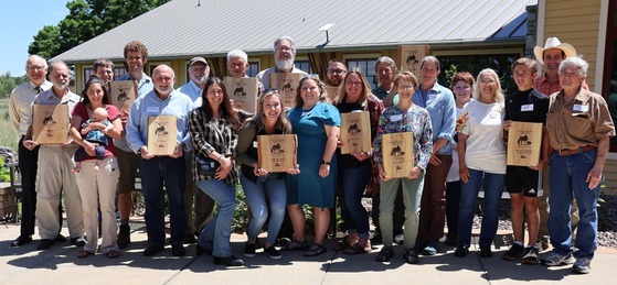 A large group of individuals pose for a picture outdoors holding wooden recognition plaques.