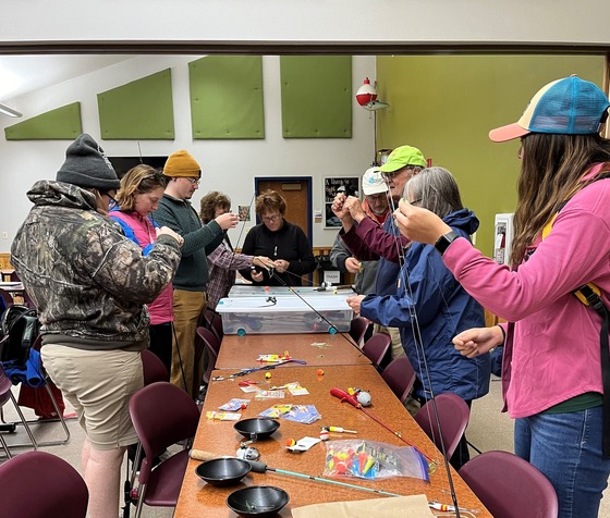 A group of people standing around a table while learning to tie fishing knots and use basic fishing equipment. 