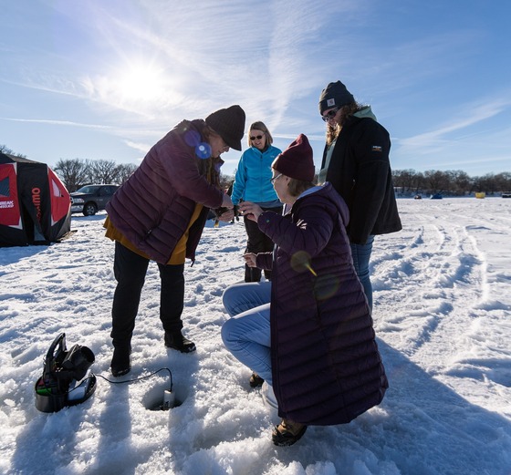 A group of individuals wearing winter jackets and hats stand around a hole in the ice and take a fish off of a hook.