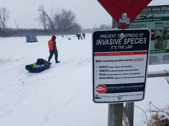 A sign with the words "Stop The Spread Of Aquatic Invasive Species" with ice anglers fishing in the background.