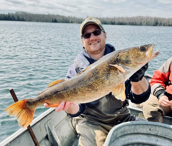 A man in a baseball cap and a inflatable life jacket holds up a large walleye in a small boat.