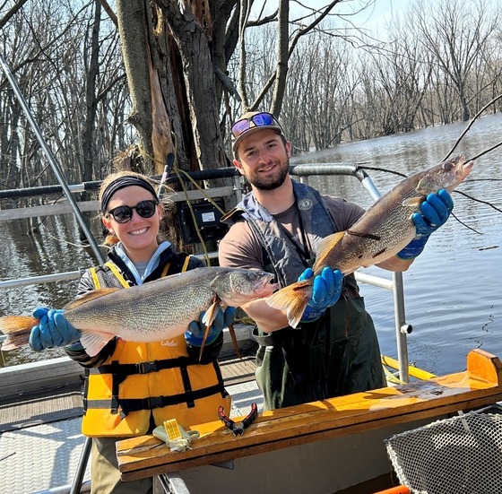 Two individuals wear life jackets stand on a boat holding up large walleye.