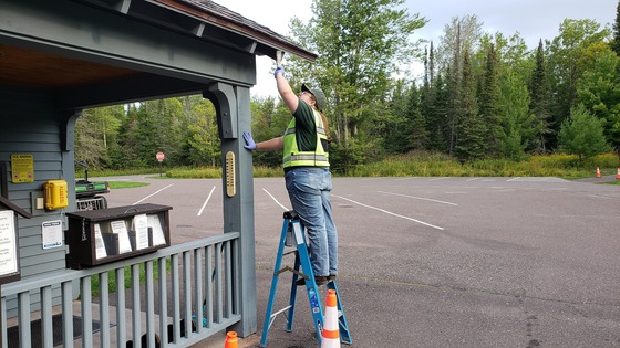 A person wearing a fluorescent vest stands on a ladder painting a wooden awning.