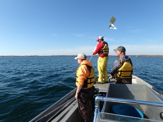 Three individuals in life jackets stand on a metal boat on a calm lake, conducting a fish survey.