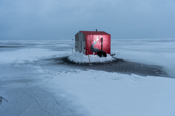 A red metal shack sits on a the ice of a frozen lake with snow piled against its base.