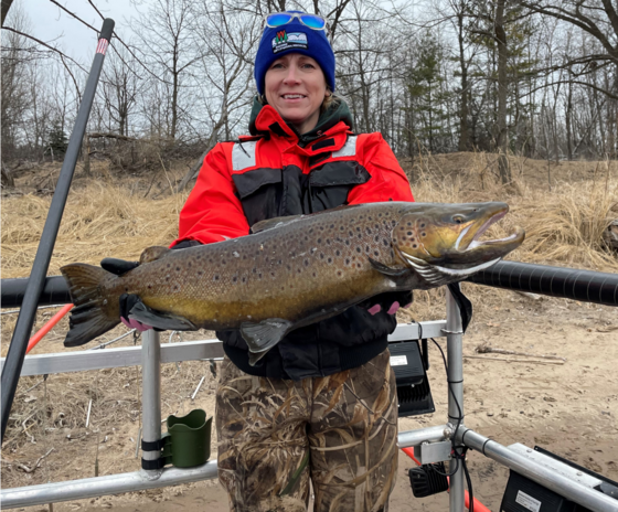 An individual in a bright orange jacket and blue hat holds up a large brown trout for a photo.