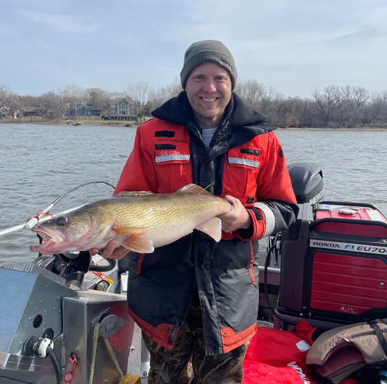 A man dressed in cold-weather gear stands on a boat holding up a large walleye with a tag in it.