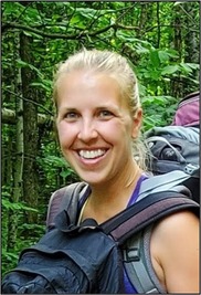 An individual with two backpacks smiles in a forest. 