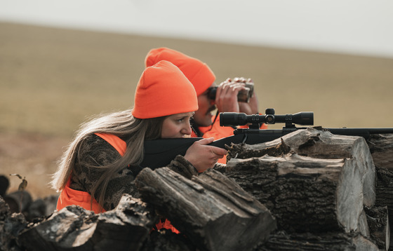 a male and female hunter in blaze orange and camouflage kneeling behind a wood pile while the woman aims her rifle at a deer out of frame.