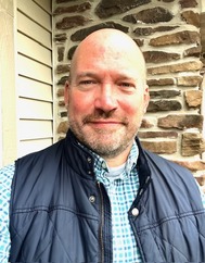An selfie of an individual in a vest in front of a brick wall outside. 