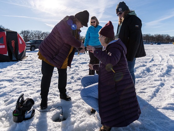 Four women sitting and standing around an ice fishing hole.