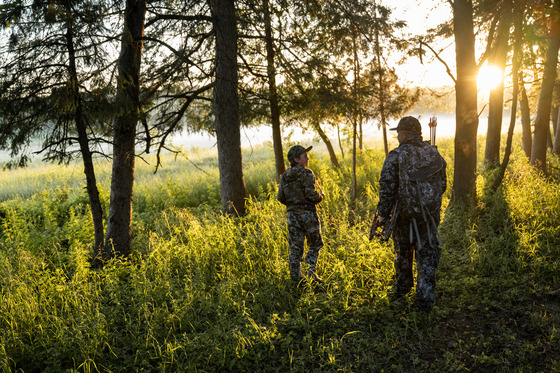 a young boy and man holding a bow in camo walking in a forest