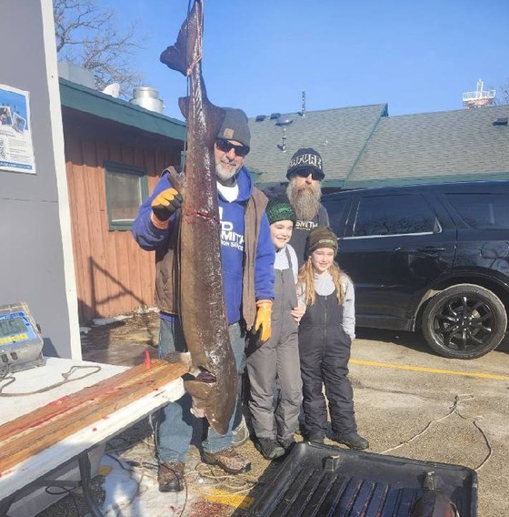 A group of four spearers stands next to a lake sturgeon hanging from its tail at a DNR registration station. 