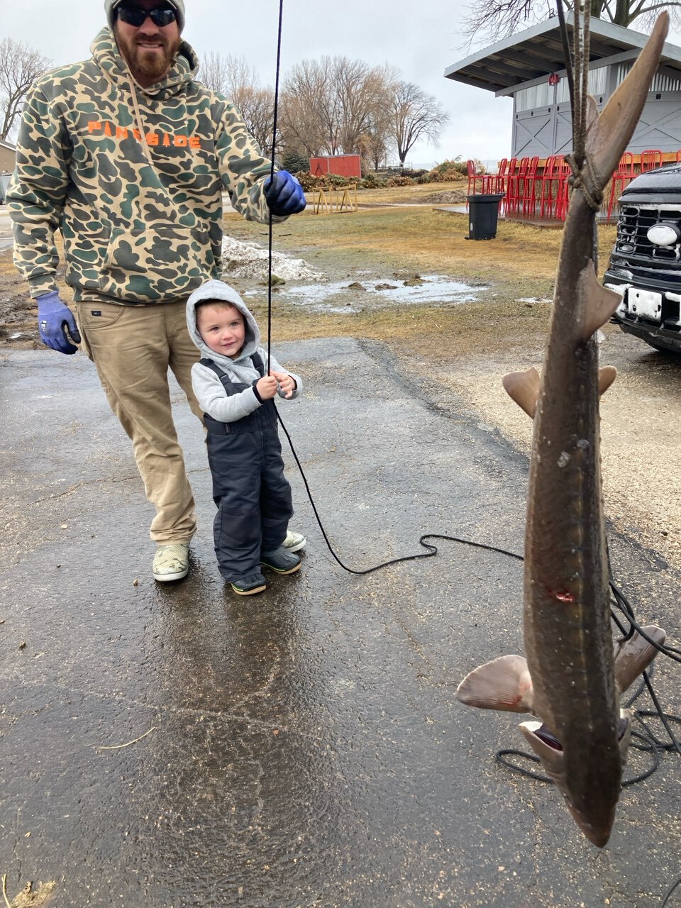 A father and son stand outdoors holding a rope as a lake sturgeon hangs from a scale. 