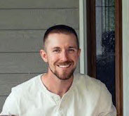 A close-up photo of a man with close-cropped hair and a big smile wearing a white shirt. 
