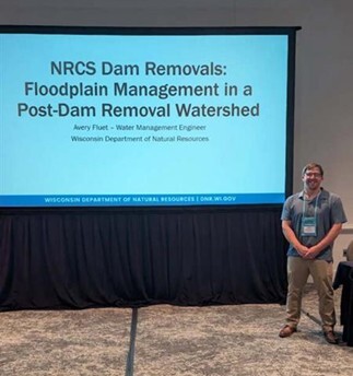 Avery Fluet standing in front of a presentation screen that says NRCS Dam Removals: Floodplain Management in a Post-Dam Removal Watershed. 