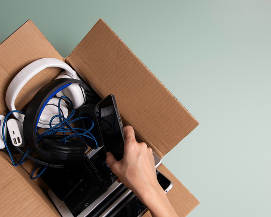 Various electronics, including headphones and a cellphone, in a cardboard box. 